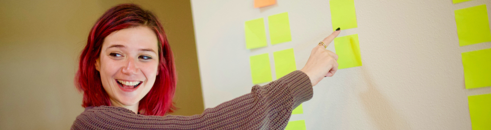 Mulher branca de cabelos avermelhados está sorridente e apontando para post its colados em um quadro branco