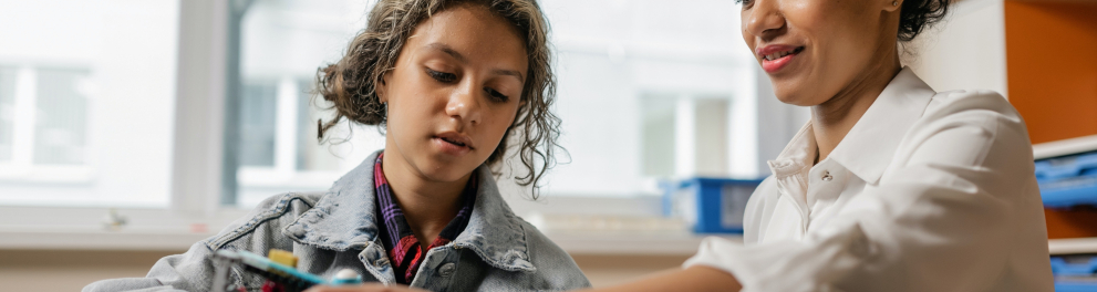 Uma menina está estudando algo com uma adulta ao seu lado, que parece ser professora