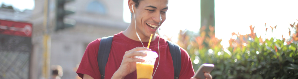 Um rapaz magro está sorridente olhando para o celular que segura com a mão esquerda, enquanto está de fones de ouvido e segurando um suco de laranja com a mão direita