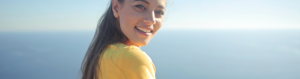 Uma linda mulher de olhos claros e cabelos castanhos compridos está sentada de costas para quem bateu a foto, porém está virada olhando para a foto e sorrindo. Ao fundo está uma vista belíssima de um mar e um céu limpo e azul