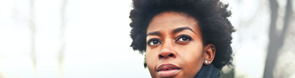 Uma linda mulher negra de cabelo black power está ao ar livre olhando para um ponto específico que não é a câmera. Ela está de casaco e cachecol