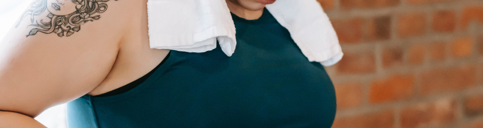 Uma mulher está com roupas de praticar exercícios e uma toalha branca em volta do pescoço. Ela está olhando para sua própria barriga enquanto aperta as gorduras laterais da região