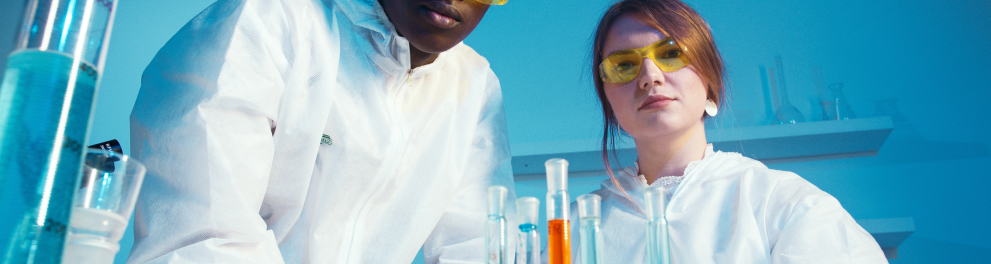 Dois cientistas de óculos de proteção e jaleco branco estão olhando sérios para quem bateu a foto. 