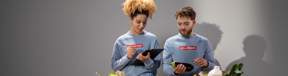 Um homem e uma mulher estão com blusas de voluntários e estão com duas caixas de papelão à frente repletas de alimentos. O homem está segurando uma berinjela e a mulher está fazendo anotações em uma prancheta