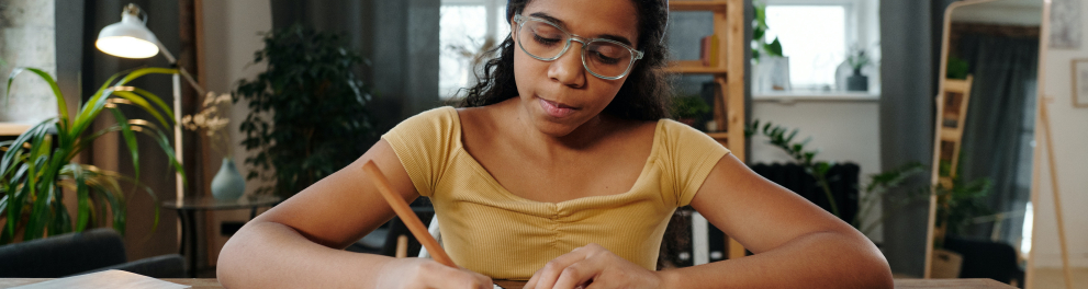 Menina escrevendo algo em um caderno apoiado sobre uma mesa dentro de casa. Ela está de óculos de grau e uma bandana azul