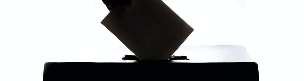 Imagem de uma mão humana inserindo um papel em uma urna de votação. Só que a imagem mostra apenas as silhuetas dos objetos