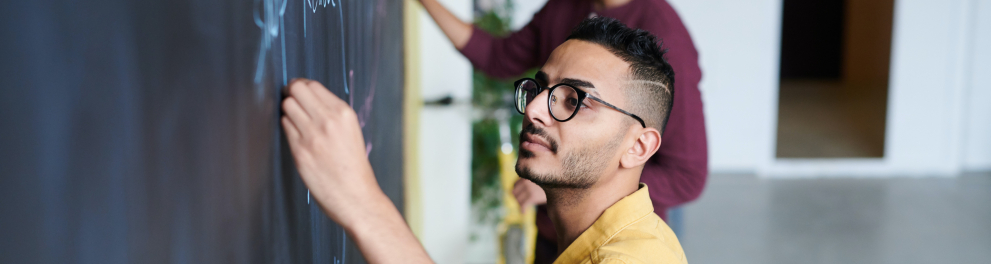 Dois jovens rapazes estão escrevendo algo em um quadro negro. Ambos estão com giz branco, óculos de grau e posicionados de perfil para quem bateu a foto