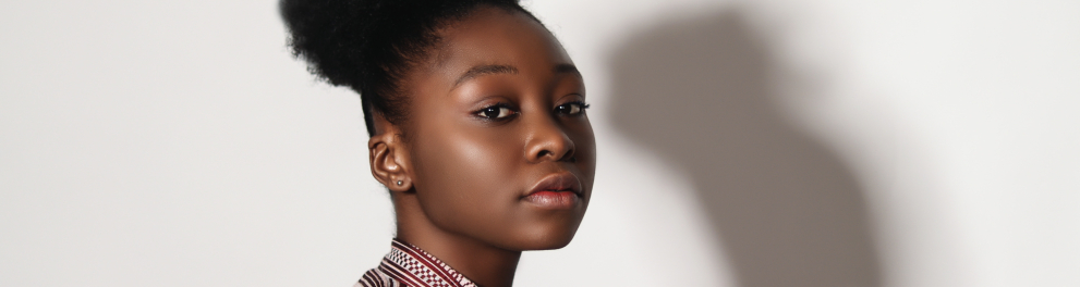 Uma jovem negra posando para a foto. No fundo, aparece a sombra dela em uma parede branca.