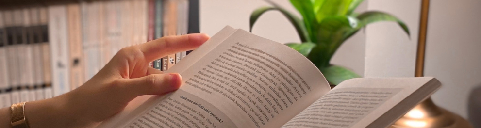 Imagem em close nas mãos de uma mulher segurando um livro aberto em frente à uma mesinha de centro, com um abajur e uma xícara de café