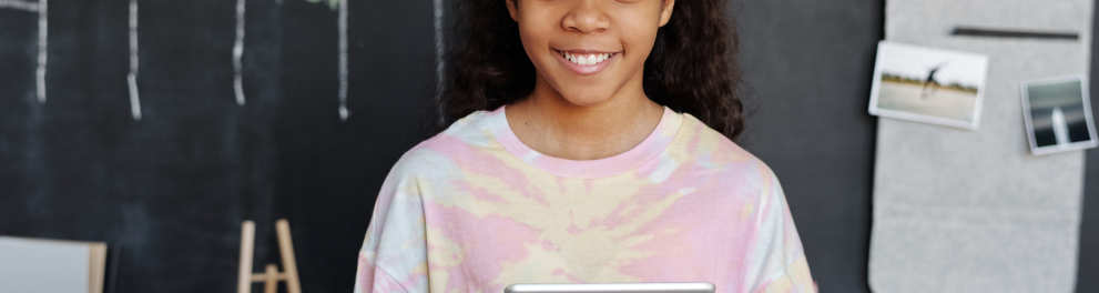 Menina negra de cabelos cacheados e camisa rosa está sorrindo para quem bateu a foto e segurando um tablet com as duas mãos