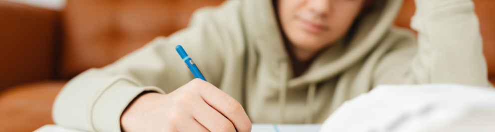 menino escrevendo algo em um caderno com uma caneta azul. Ele está de frente pra quem bateu a foto e com camisa de mangas compridas e capuz