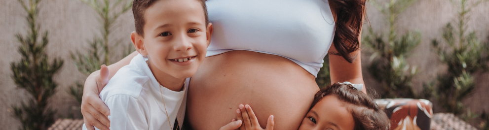 Duas crianças estão tocando na barriga da mãe grávida, que está no centro da imagem. As crianças estão olhando sorridentes para quem bateu a foto 