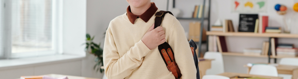 Menino em sala de aula está em pé, com a mochila nas costas, segurando uma das alças e sorrindo sem mostrar os dentes para quem bateu a foto