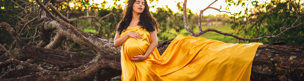 Mulher grávida usando um longo vestido amarelo está posando para a foto encostada em um tronco de árvore caído em meio à uma vegetação