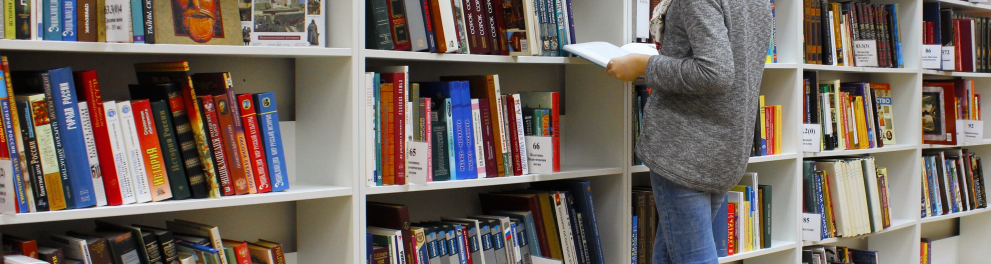 Uma mulher está em uma biblioteca de costas para quem bateu a foto. Ela está com um livro em mãos