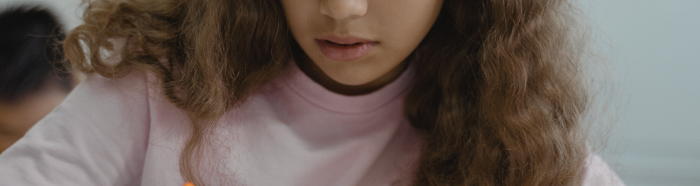 Menina está em sala de aula escrevendo algo em seu caderno. Ela está de camisa rosa de mangas compridas e cabelo castanho solto e longo