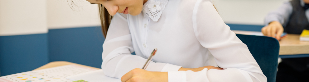 Menina está em sala de aula escrevendo algo em seu caderno. Ela está de camisa branca de mangas longas e tem cabelos lisos e castanhos compridos