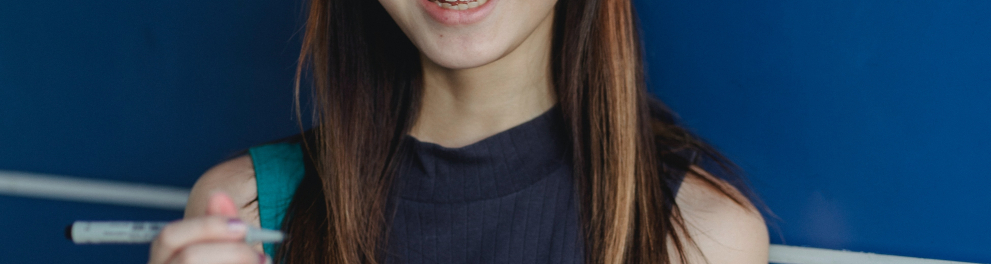 Adolescente asiática está segurando livros com a mão esquerda e uma caneta com a mão direita. Ela está olhando sorridente para quem bateu a foto