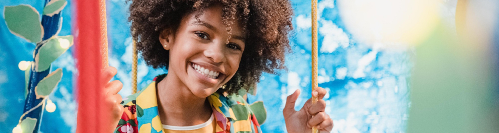 Menina negra de cabelo black power está sorridente, sentada em um balanço e posando para quem bateu a foto. Ao fundo, notamos um cenário bem colorido, com a cor azul clara predominando
