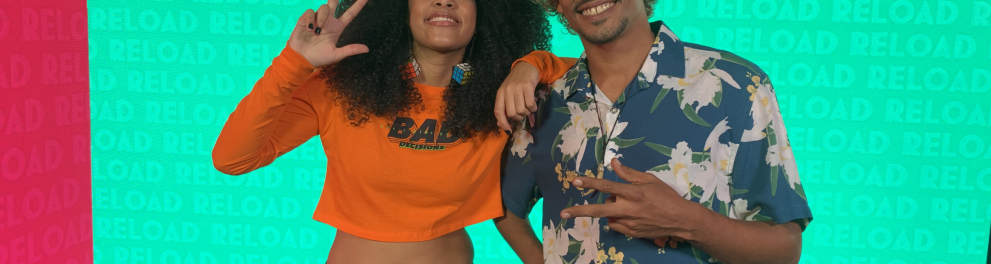 Os dois apresentadores da série Reload estão lado a lado posando sorridentes para a foto. É um rapaz e uma moça negros. Acima deles está a palavra "reload"