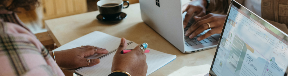 Close em uma mesa de estudo, com notebook aberto e as mãos de uma mulher sobre um caderno