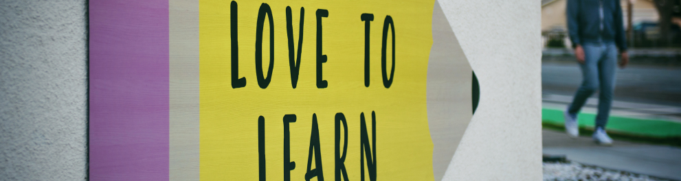 Uma placa no formato de lápis e nas cores amarela e roxa está colada em um muro branco e tem a seguinte frase na parte amarela: "Love to learn"