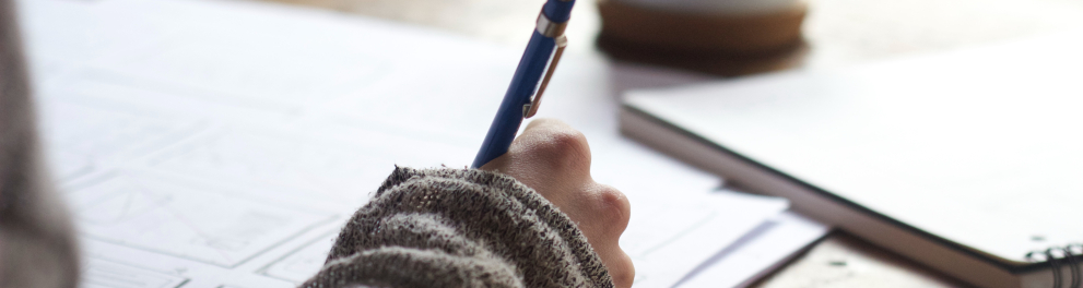 Imagem em close de uma mão direita branca segurando uma lapiseira azul e escrevendo algo em um caderno