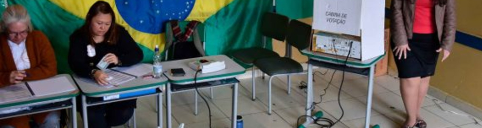 A imagem mostra mesários e uma pessoa votando em uma eleição brasileira