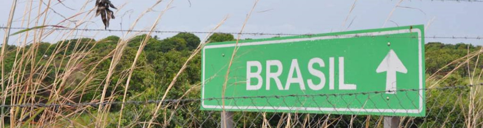 Uma placa verde posicionada à beira de uma estrada com a palavra "Brasil" escrita em branco e uma seta  apontada para cima ao lado da palavra