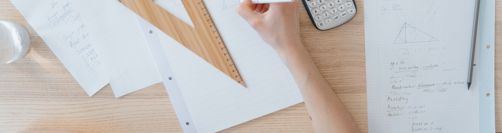 Imagem mostra uma Mão escrevendo sobre papel, com régua e calculadora próximos