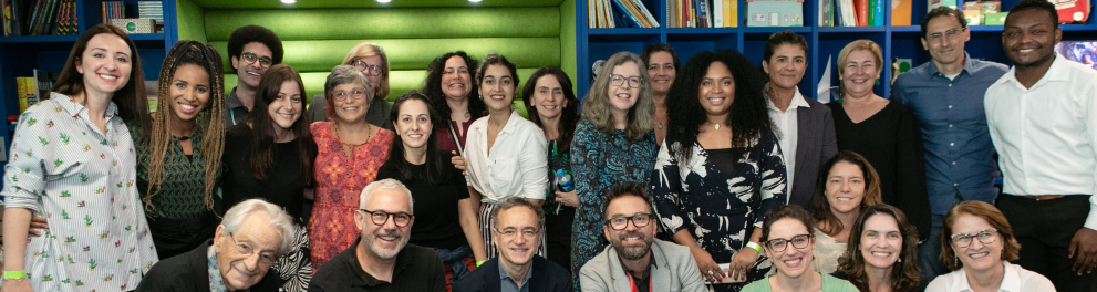 Diversas pessoas estão de pé e agachadas (as de pé, atrás das que estão agachadas) em uma sala. Todas sorriem.