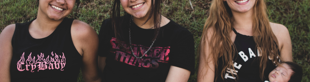 Três jovens mulheres estão sentadas lado a lado e sorrindo para quem bateu a foto. A da esquerda e a do centro são morenas e parecem ser irmãs gêmeas, já a da direita é loira, branquinha e está com um bebê no colo