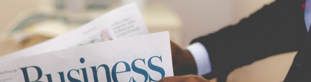 Zoom nas mãos de um homem negro trajando paletó e relógio de gala. Ele está segurando um jornal que fala sobre negócios e finanças