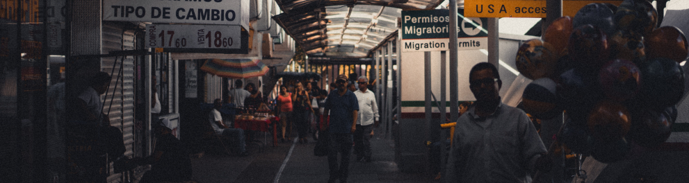 Imagem de um corredor escuro com pessoas andando ou paradas nele. Ao lado esquerdo tem coisas escritas em espanhol e ao lado direito em inglês