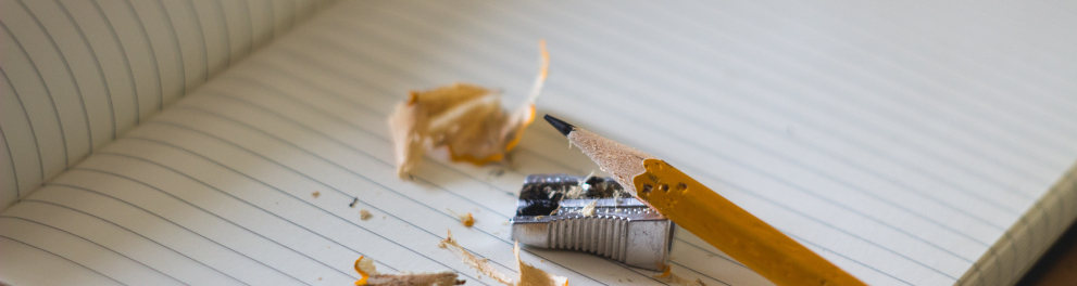Um lápis amarelo acabou de ser apontado e está em cima de um caderno aberto com as raspas ao redor e o apontador