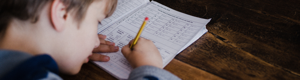 Um menino sentado escrevendo algo em seu caderno. Ele está de costas para quem bateu a foto, mas podemos ver bem o caderno com várias equações de matemática