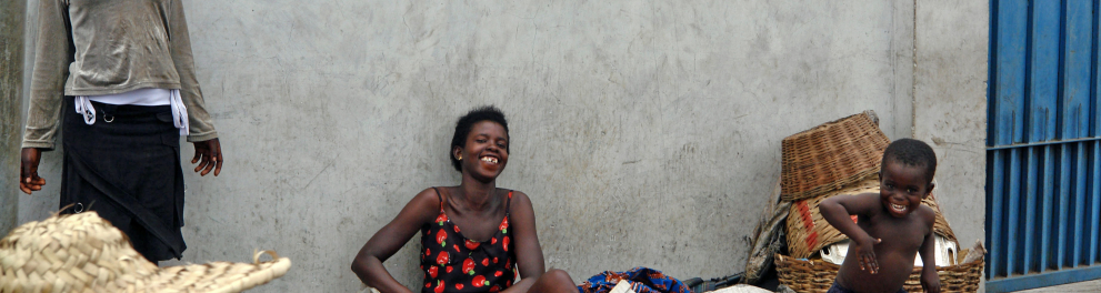 Uma mulher africana está sentada no chão de uma rua encostada à parede de uma casa, enquanto uma criança está em pé ao seu lado sorridente e dançando. 