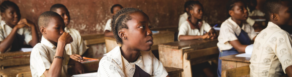 O interior de uma sala de aula africana. Na imagem vemos algumas crianças negras de uniforme escolar sentadas em suas cadeiras e olhando todas em uma mesma direção, que certamente é ao professor e o quadro.