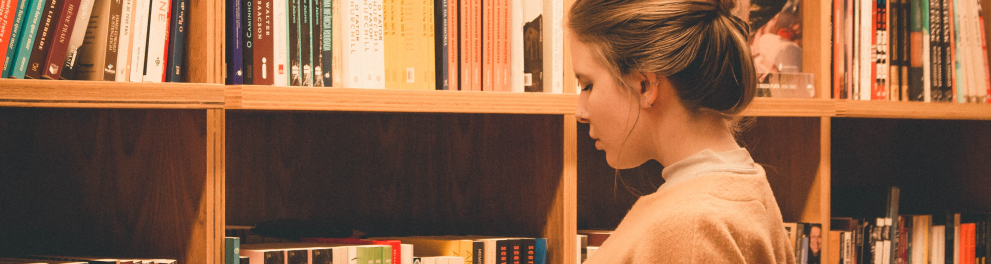 Uma mulher está em pé em uma biblioteca, com um livro na mão esquerda e olhando para uma prateleira com outros livros