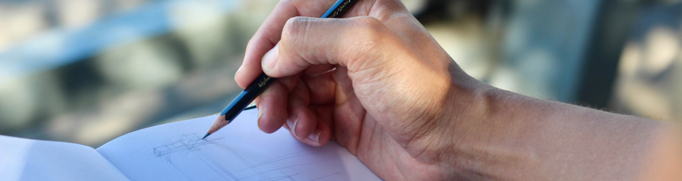 Mão direita de uma pessoa está segurando um lápis e prestes a escrever em um caderno ao ar livre