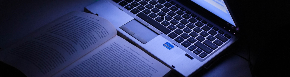 Imagem de um notebook aberto e ligado em cima de uma mesa, com um livro aberto e um celular ao seu lado. Os objetos estão em um ambiente escuro e a iluminação é apenas da tela do notebook