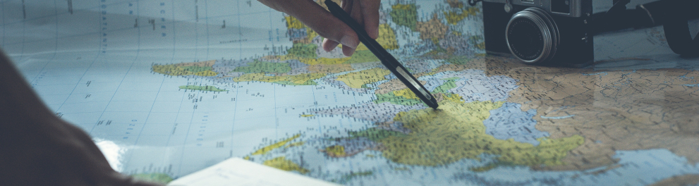 Mãos de uma pessoas estão em destaque na foto. A mão esquerda está segurando uma caneta apoiada em um mapa de papel e a mão direita está em um livro aberto abaixo do mapa
