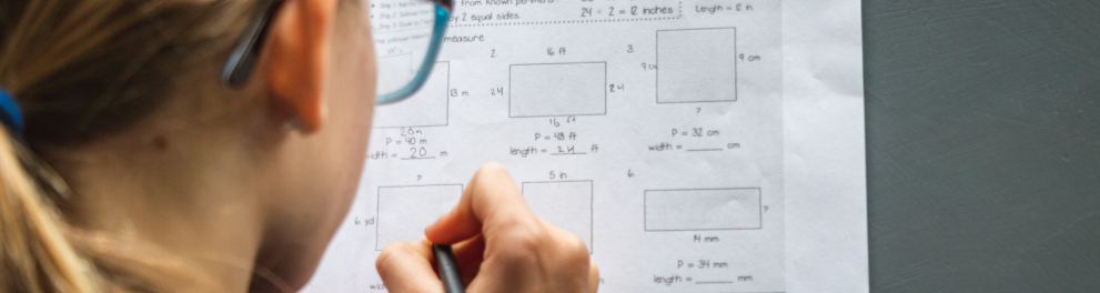 Imagem de uma menina loira realizando uma prova de matemática. A imagem foi feita por trás da menina, então podemos ver a sua prova