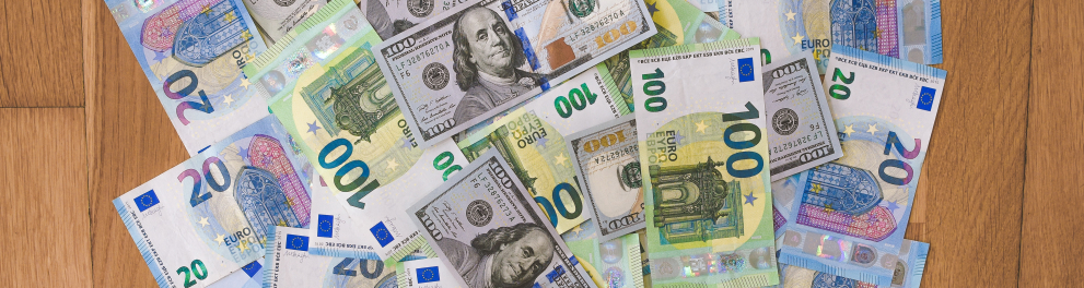 Várias cédulas de dólares e euros estão reunidas e amontoadas sobre uma superfície de madeira