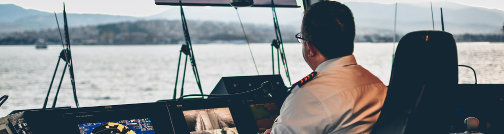 Um homem de costas para quem bateu a foto está sentado com uniforme de capitão e parece estar comandando um painel de navio