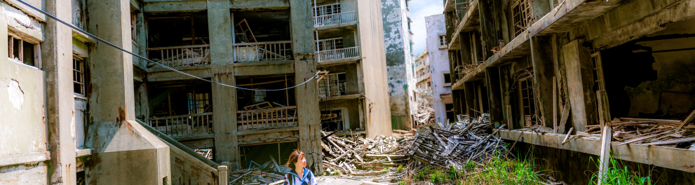 Uma jovem oriental está parada no centro da imagem olhando ao seu redor, que são apenas ruínas e muito desastre com prédios demolidos e muito concreto e poeira