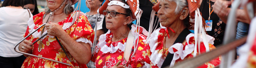 Três senhoras com roupas típicas de Pernambuco e chapéus de cangaceiras estão em algum tipo de apresentação de dança ao ar livre