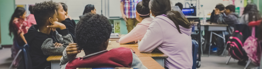 Uma sala de aula com alunos olhando atentamente para o professor em pé falando algo