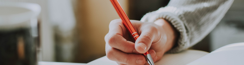 Mão direita de uma de uma pessoa segurando uma caneta que está prestes a escrever em um caderno