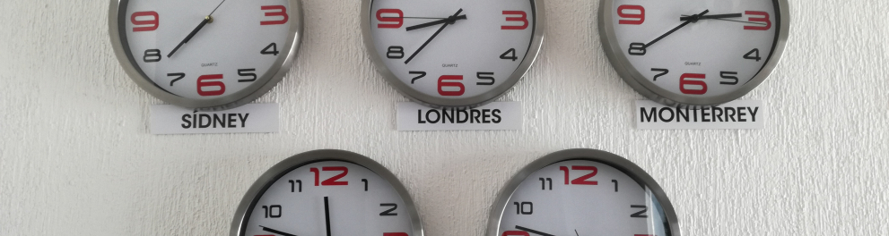 Cinco relógios redondos de parede estão lado a lado marcando horários diferentes e cada um corresponde a uma cidade do mundo, pois há plaquinhas com os nomes das cidades abaixo de cada relógio